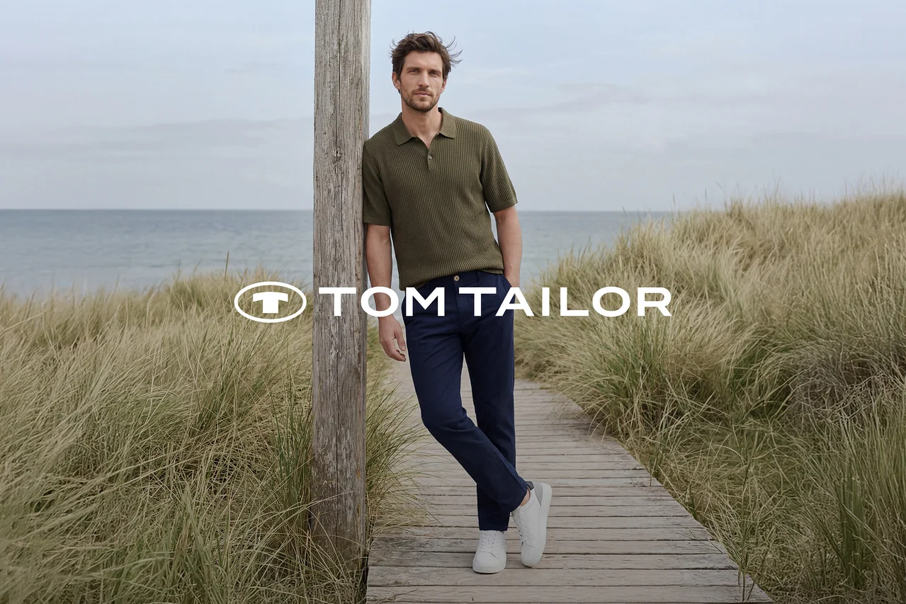 Mężczyzna w zielonej koszulce polo, granatowych spodniach i białych sneakersach opiera się o drewniany słup na pomoście przy morzu, otoczony wysoką trawą. Logo TOM TAILOR.