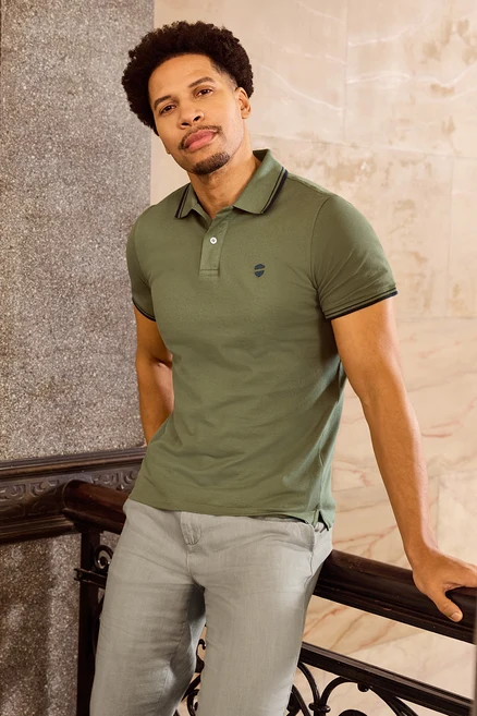Mężczyzna opiera się o barierkę, ubrany w khaki koszulkę polo o splocie pique.