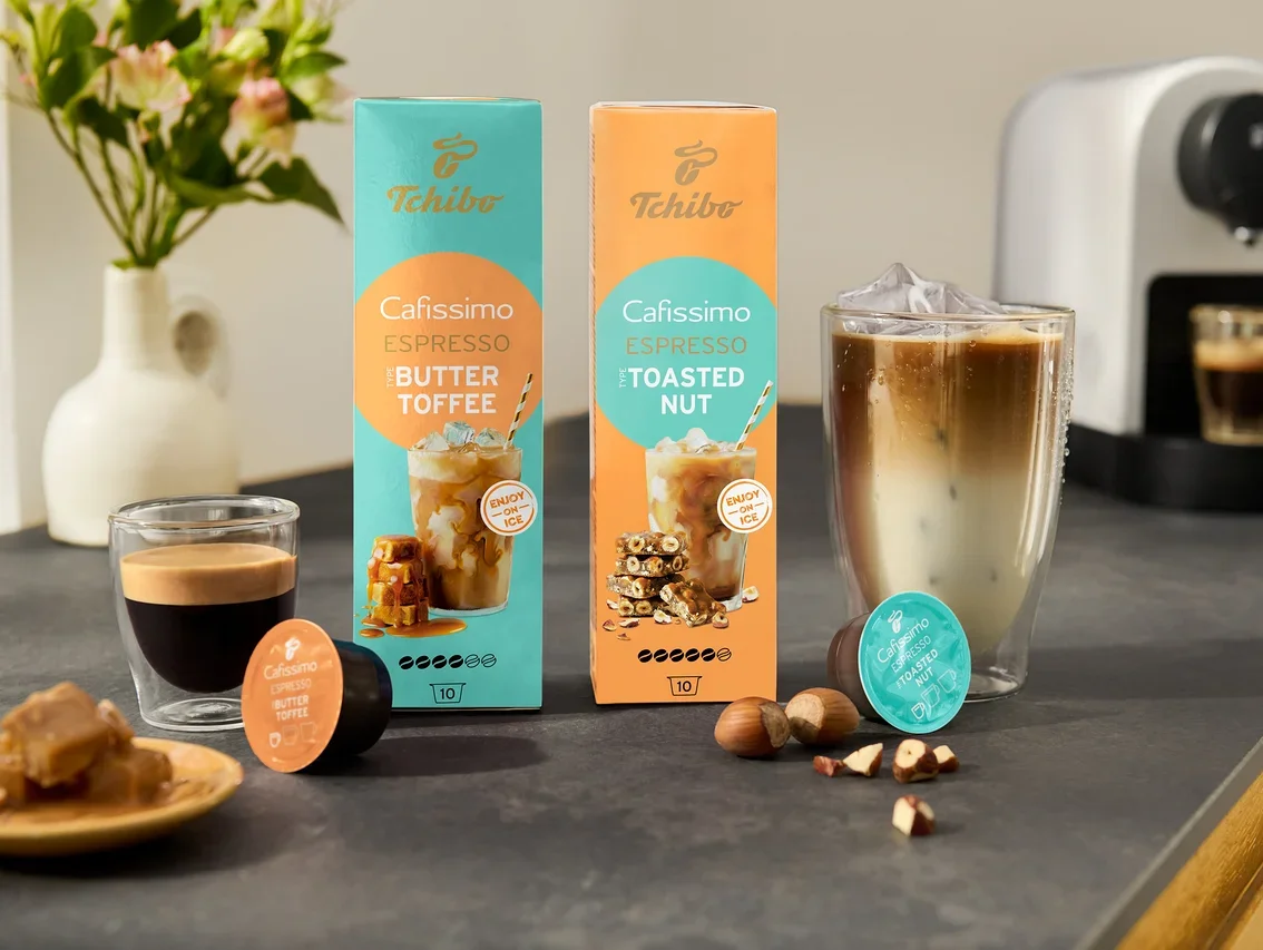 A konyhapulton két csomag Cafissimo Flavoured Espresso áll, egy türkiz színű Buttertoffee, egy narancssárga Toasted Nut ízesítésű, mellette egy fehér Cafissimo pure+ gép.
