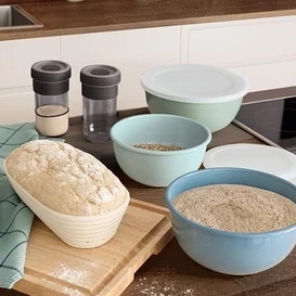 Na drewnianym blacie kuchennym leżą drewniana deska do krojenia, koszyk rozrostowy z chlebem, dwie przyprawniki i trzy miski.