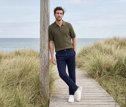 Mężczyzna w oliwkowej koszulce polo, granatowych spodniach i białych sneakersach opiera się o drewniany słupek na tle plaży i morza.