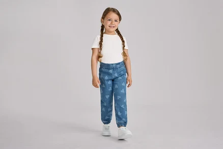 Uśmiechnięta dziewczynka z dwoma warkoczami stoi, ubrana w biały T-shirt, niebieskie spodnie na gumce – fason „Ella” w serduszka i białe trampki.