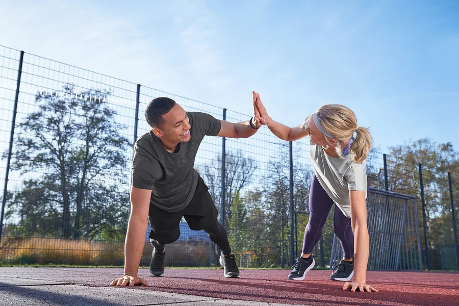 Mężczyzna w koszulce sportowej, jasnoszarej nakrapianej i szortach sportowych oraz kobieta w legginsach i funkcyjnej koszulce Klima-Tec przybijają sobie piątki podczas ćwiczeń na zewnątrz.