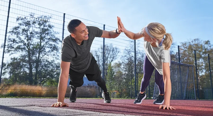 Mężczyzna w koszulce sportowej, jasnoszarej nakrapianej i szortach sportowych oraz kobieta w legginsach i funkcyjnej koszulce Klima-Tec przybijają sobie piątki podczas ćwiczeń na zewnątrz.