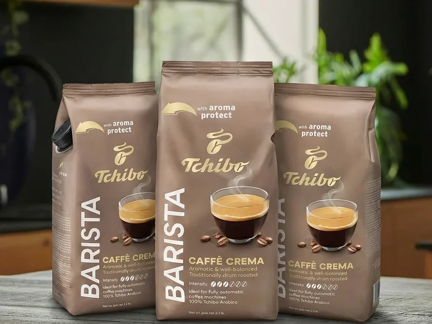 Trzy opakowania Tchibo Barista Caffè Crema na drewnianym blacie przed oknem.