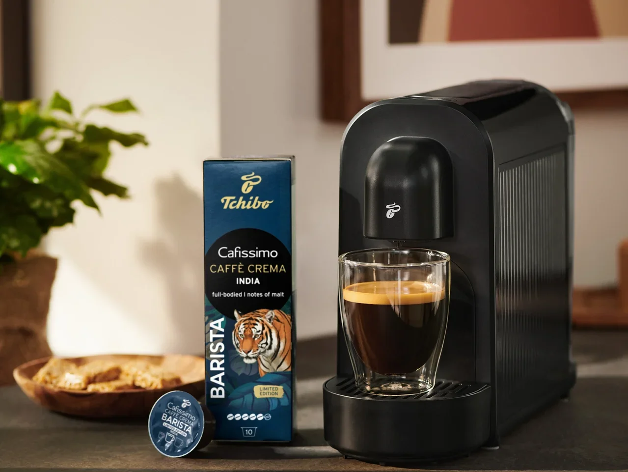 Czarny ekspres do kawy Tchibo Cafissimo ze szklanką kawy, obok opakowanie kapsułek Tchibo Cafissimo Caffè Crema India i jedna kapsułka.