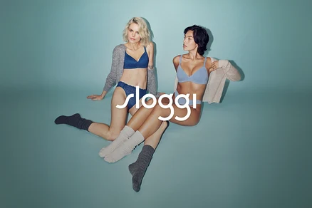 Dwie kobiety siedzą na turkusowym tle. Blondynka ma na sobie niebieski biustonosz, wzorzyste majtki, szary kardigan i ciemne skarpetki. Ciemnowłosa kobieta nosi jasnoniebieski biustonosz, majtki, beżowy kardigan i jasne skarpetki. Białe logo „sloggi”.