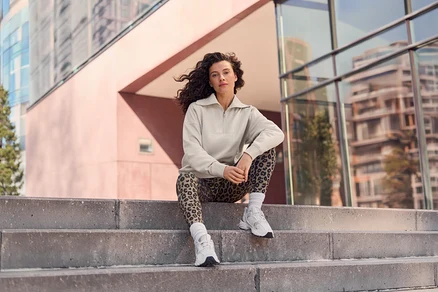 Kobieta z kręconymi włosami siedzi na betonowych schodach, ubrana w beżowy top z zamkiem błyskawicznym, legginsy w panterkę i białe sneakersy, z nowoczesnymi budynkami w tle.