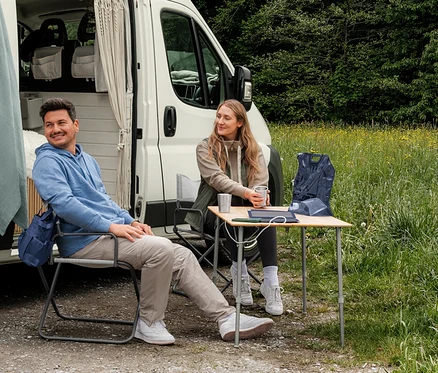 Mężczyzna w jasnoniebieskiej bluzie dresowej z kapturem i kobieta w beżowej rozpinanej bluzie dresowej i kamizelce hybrydowej khaki siedzą przed kamperem. Na stole leży granatowa kamizelka typu packable.