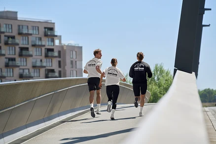 Trzy osoby biegną po moście: mężczyzna w czarnych szortach sportowych i białej koszulce sportowej, kobieta w czarnych legginsach sportowych „ActivePower” i białej koszulce sportowej, oraz kobieta w czarnej bluzie i czarnych kolarkach „ActivePower”.