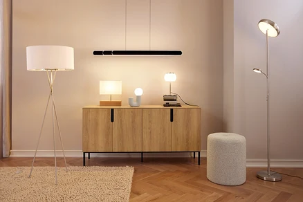 Nowoczesny pokój z drewnianą komodą, na której stoją trzy lampy i przedmiot dekoracyjny, pod czarną liniową lampą wiszącą. Po lewej stronie na dywanie stoi biała lampa podłogowa na trójnogu, a po prawej srebrna lampa podłogowa z oświetleniem górnym i lampką do czytania obok szarego pufa.