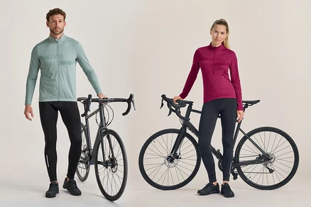 Mężczyzna i kobieta stoją obok czarnych rowerów szosowych. Mężczyzna ma na sobie jasnozieloną koszulkę rowerową z długim rękawem i czarne spodnie rowerowe. Kobieta ma na sobie ciemnoróżową koszulkę rowerową z długim rękawem i czarne spodnie rowerowe.