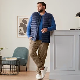 Mężczyzna opiera się o komodę, ubrany w niebieską kamizelkę pikowaną, niebieski sweter lniany, karmelowe spodnie chino i buty typu sneaker.