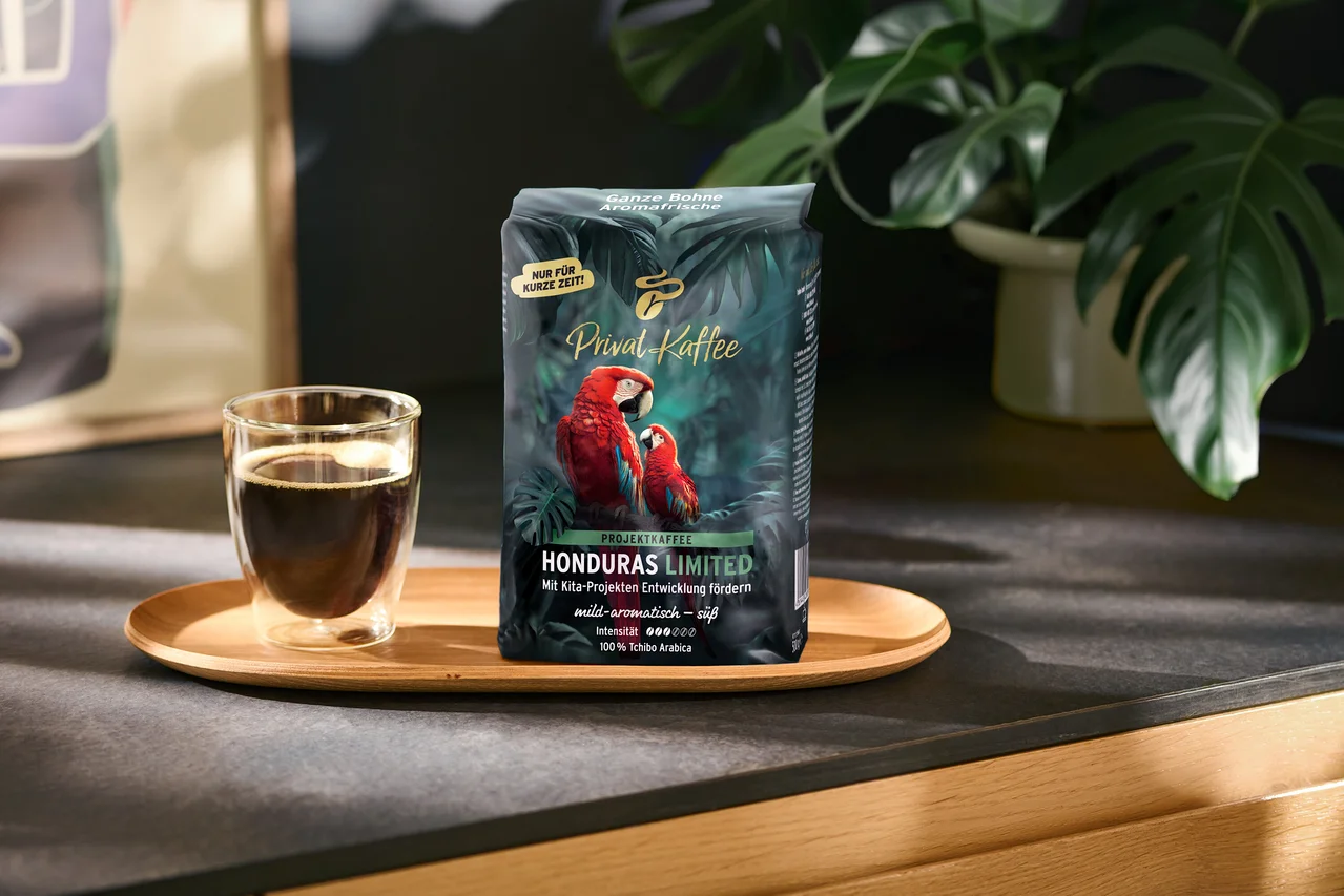 Ciemnozielona torebka kawy „Privat Kaffee Honduras Limited” z dwoma czerwonymi arami i szklana filiżanka czarnej kawy stoją na drewnianej tacy. Na torebce widnieją napisy takie jak „NUR FÜR KURZE ZEIT!” i „100% Tchibo Arabica”. W tle znajduje się zielona roślina.