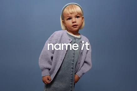 Małe dziecko z blond włosami i niebieską opaską na głowie ma na sobie dżinsowe ogrodniczki i liliowy kardigan na tle niebieskiego tła.