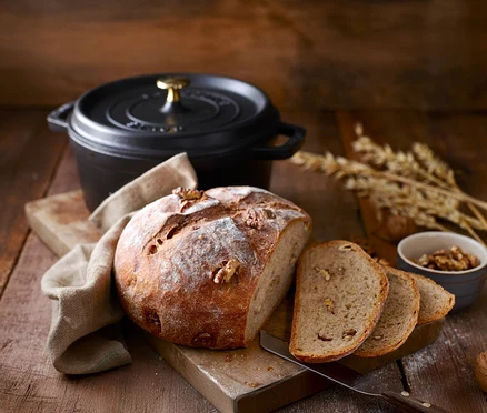 Bochenek chleba orzechowego i kromki chleba na desce do krojenia z czarnym garnkiem STAUB »Cocotte 24« w tle.