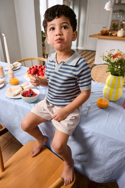 Chłopiec siedzi na krawędzi stołu z truskawką na widelcu, ubrany w Kinder-Shorts i Kinder-Strick-Poloshirt. Na stole wazon i patera na tort.