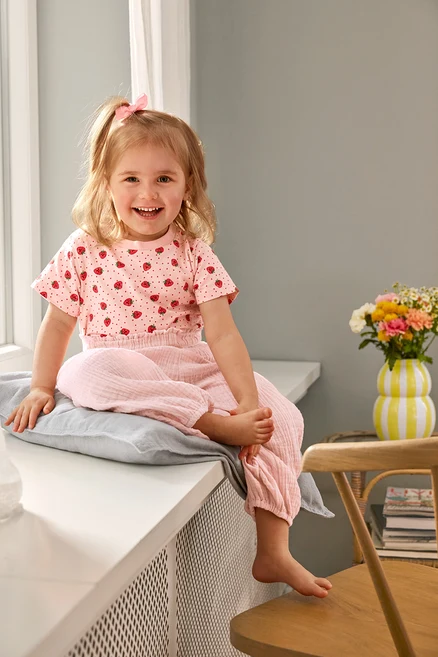 Uśmiechnięta dziewczynka siedzi w 2 Kinder-T-Shirts i Kinder-Musselin-Hose na poduszce. Na krześle stoi wazon.