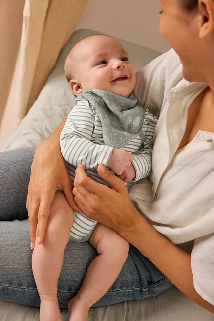 Matka trzyma uśmiechnięte dziecko ubrane w body i muślinową chustkę.