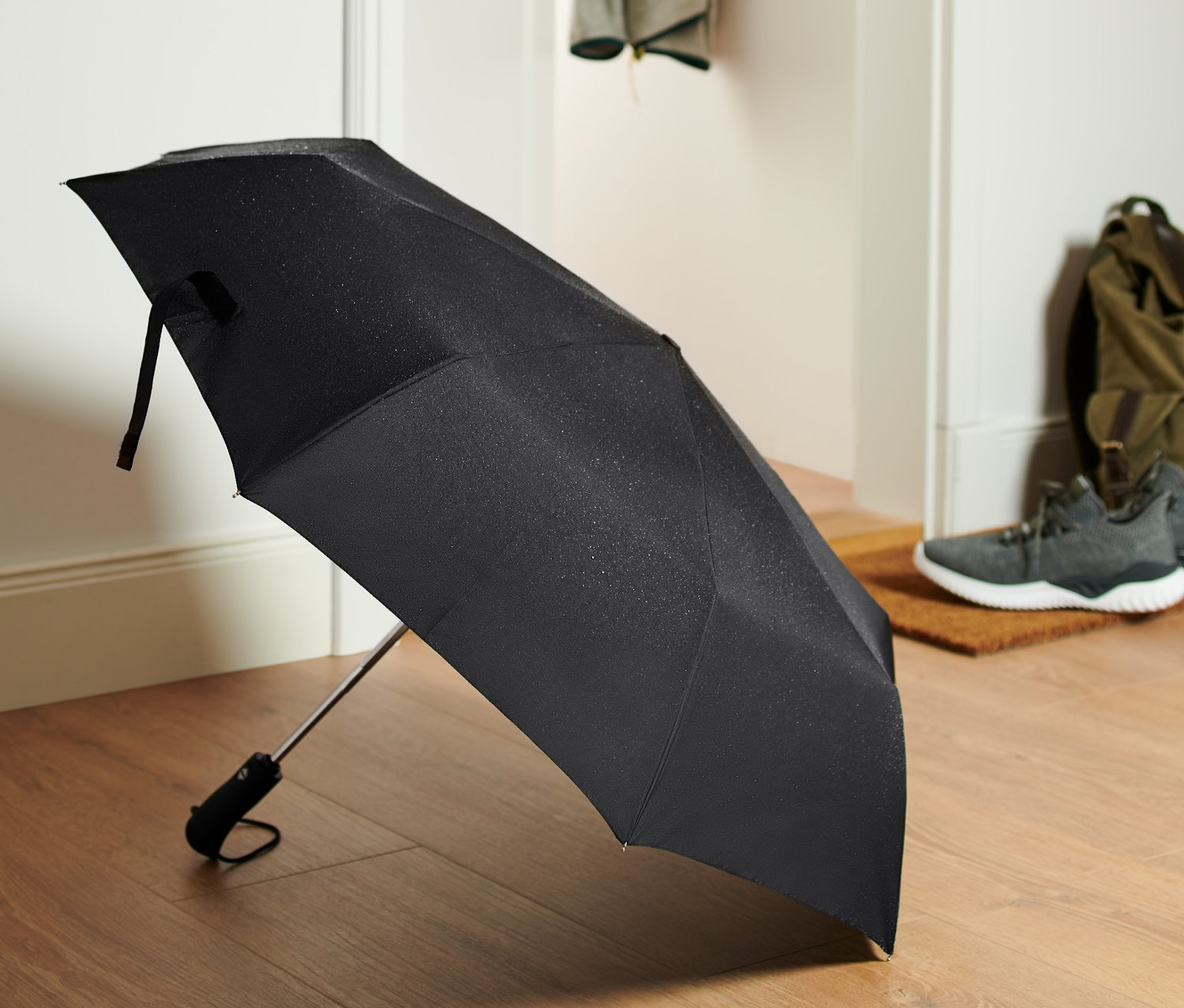 Czarny kieszonkowy parasol automatyczny leży na drewnianej podłodze przy drzwiach.