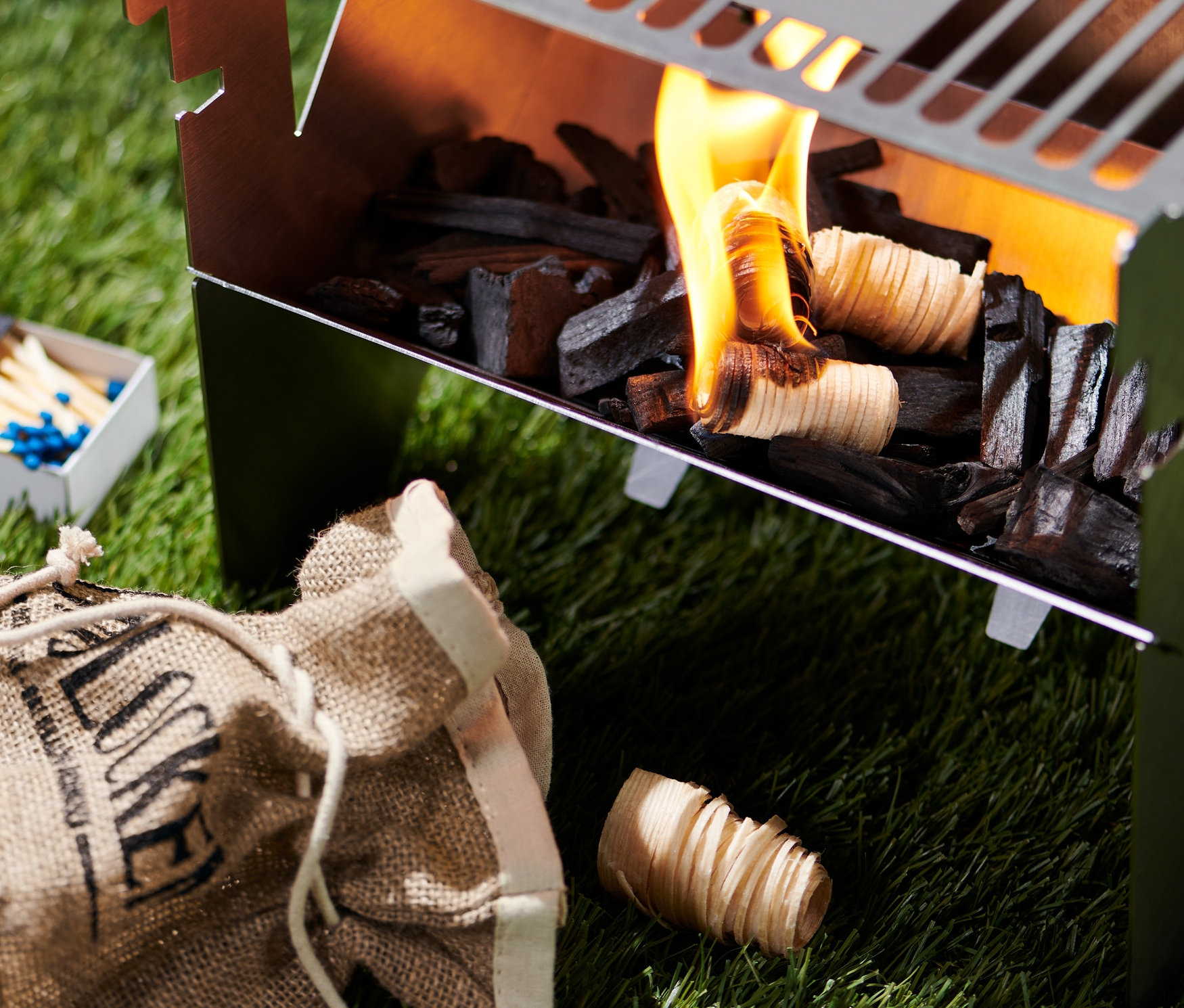 Paląca się rozpałka do węgla do grillowania w grillu. Obok grilla leżą zapałki i worek z rozpałką.