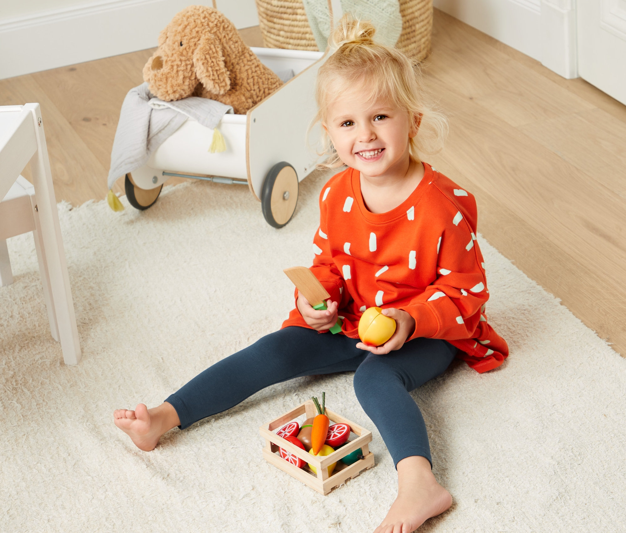 Blondwłosa dziewczynka siedzi na dywanie i bawi się drewnianymi owocami. Ma na sobie pomarańczową bluzę i legginsy z bawełny ekologicznej, 2 pary.