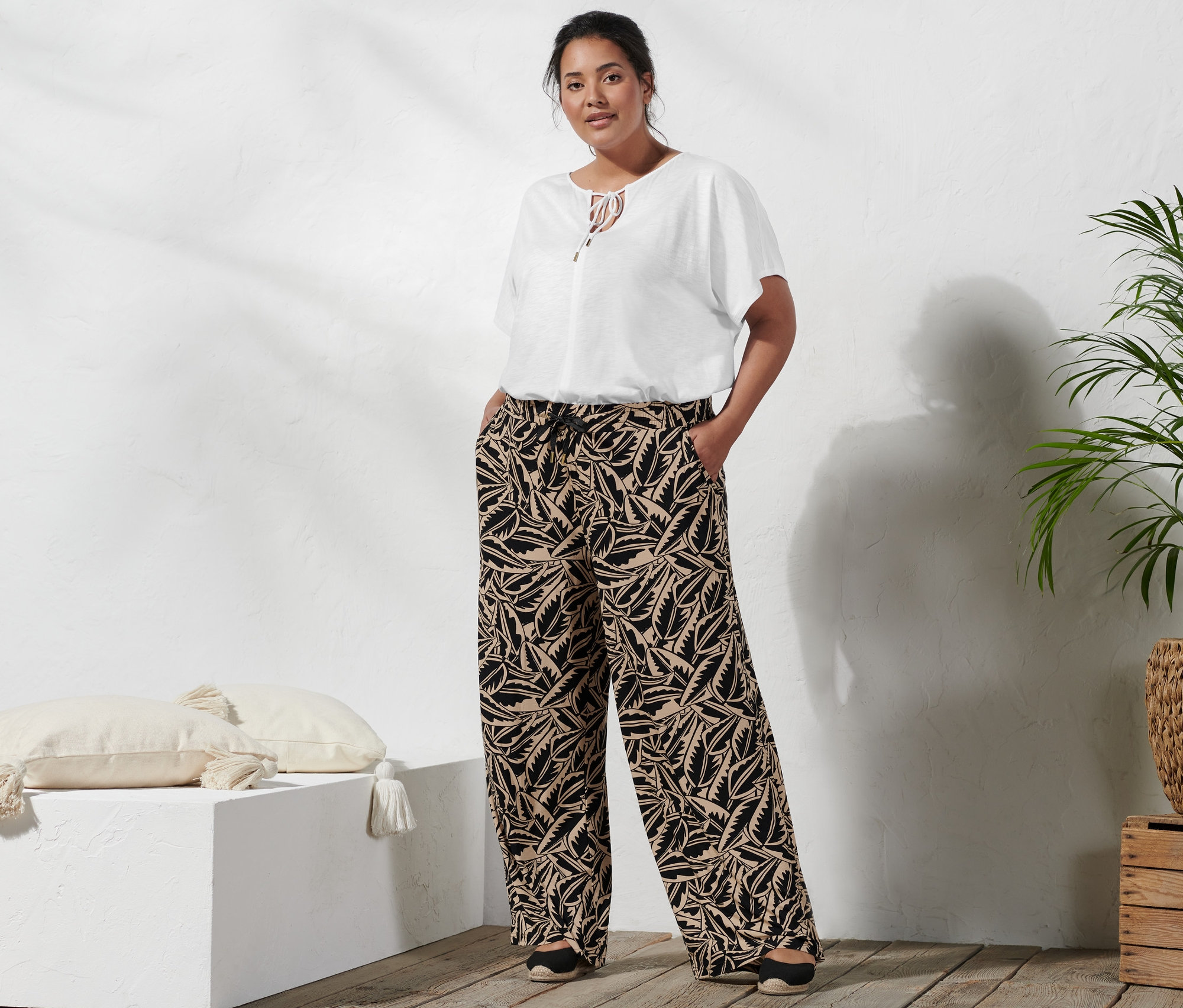 Kobieta stoi z rękami w kieszeniach spodni palazzo z nadrukiem w kolorze czarno-beżowym, ma na sobie bluzkę z wiązaniem i espadryle na koturnie.