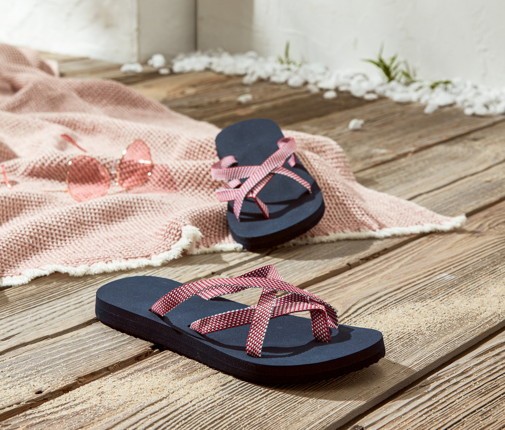 Dwie pary klapki japonki leżą na drewnianej podłodze obok różowych okularów przeciwsłonecznych i różowego koca plażowego.