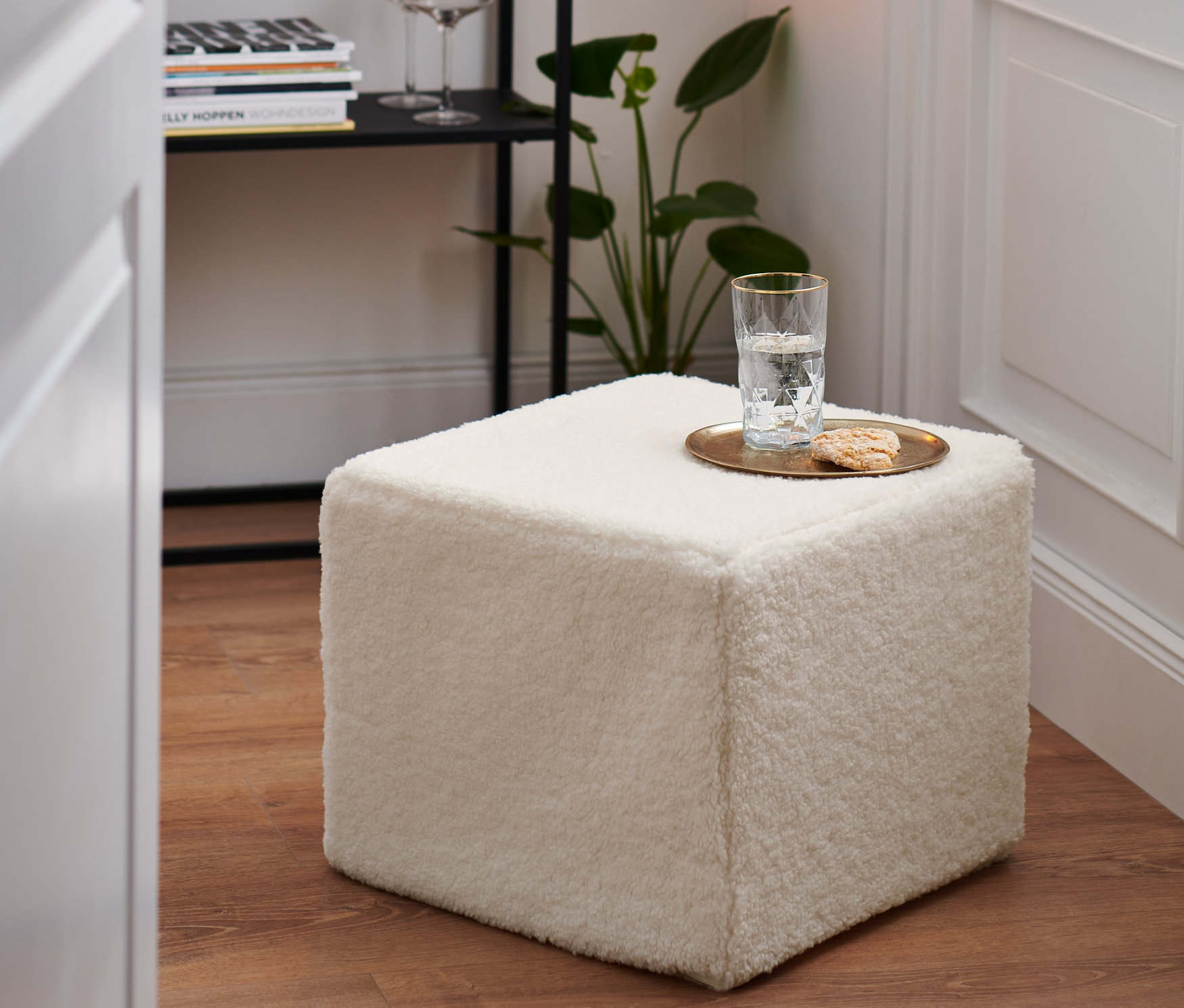 Na drewnianej podłodze stoi biała pufa. Na pufie stoi szklanka wody i ciasteczko.
