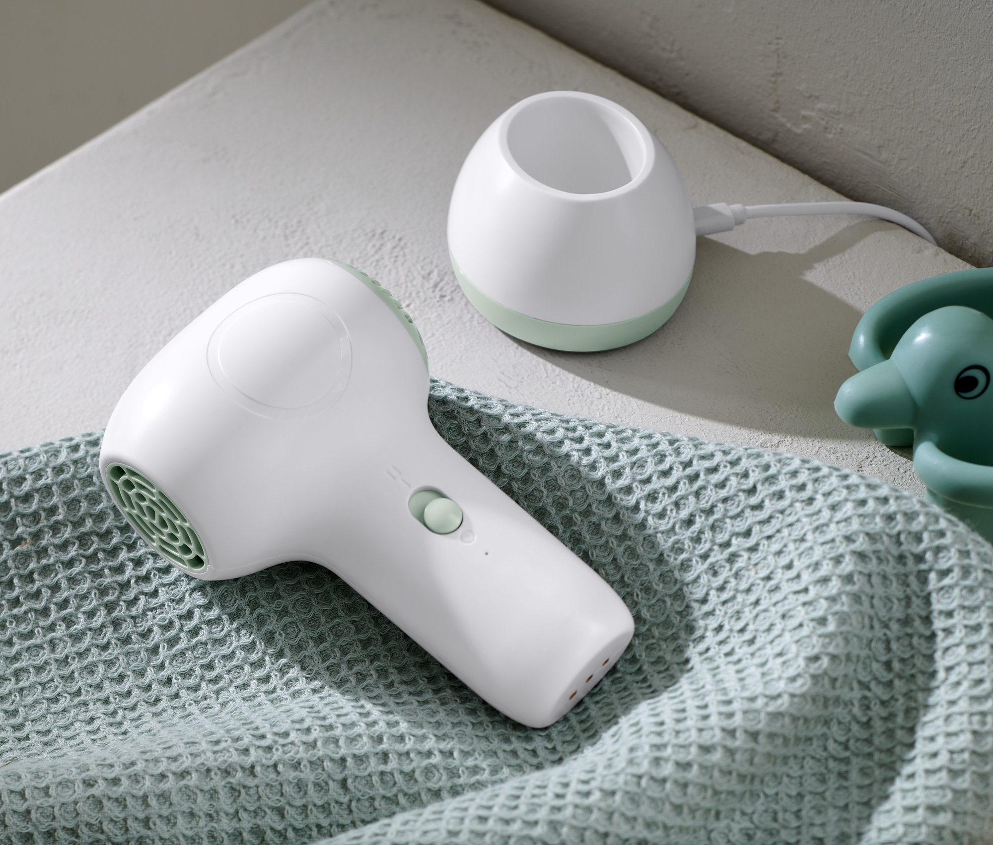 Biała akumulatorowa suszarka do włosów dla dzieci leży na jasnoniebieskim ręczniku obok stacji ładującej i zabawki do kąpieli.