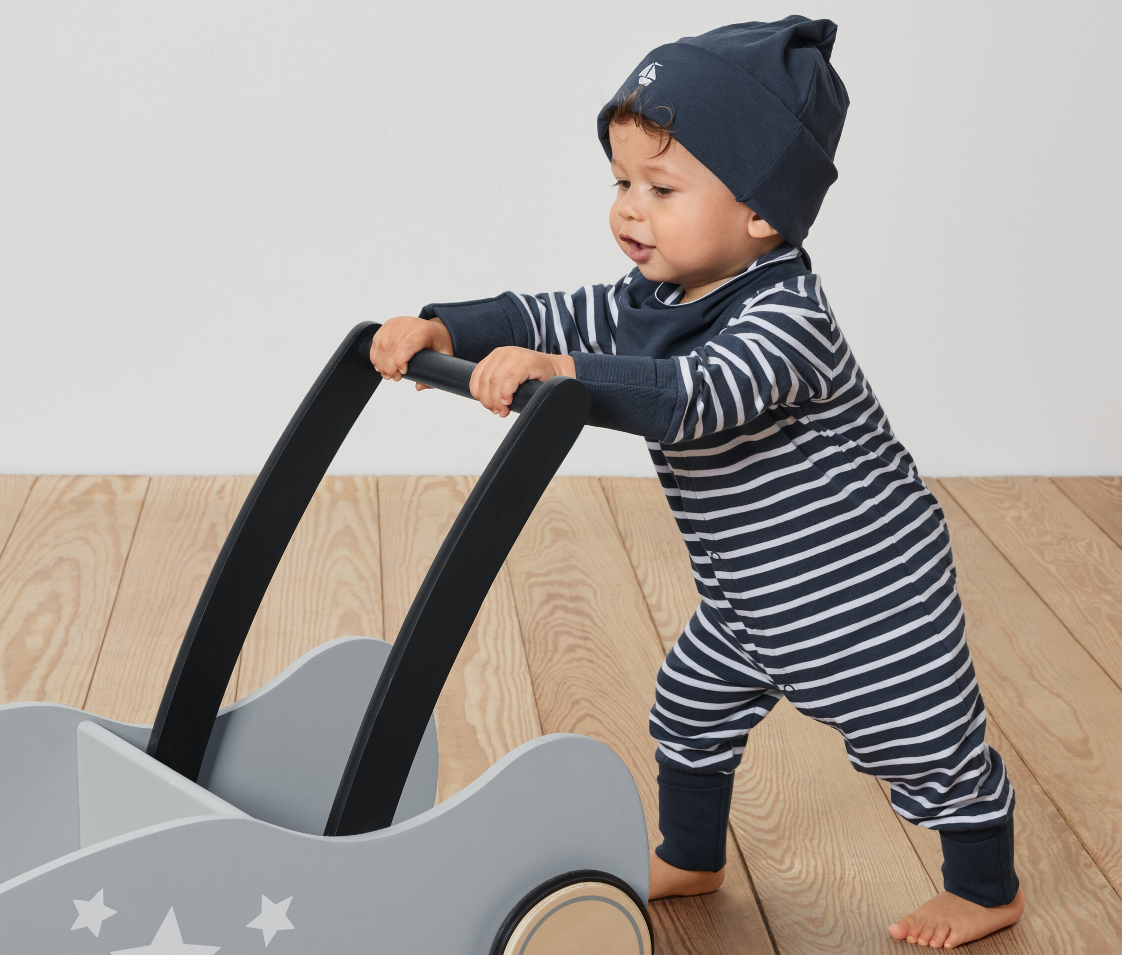 Małe dziecko pcha szary wózek z gwiazdami. Ubrane jest w 3-częściowy zestaw niemowlęcy: kombinezon w paski, czapkę i śliniaczek.