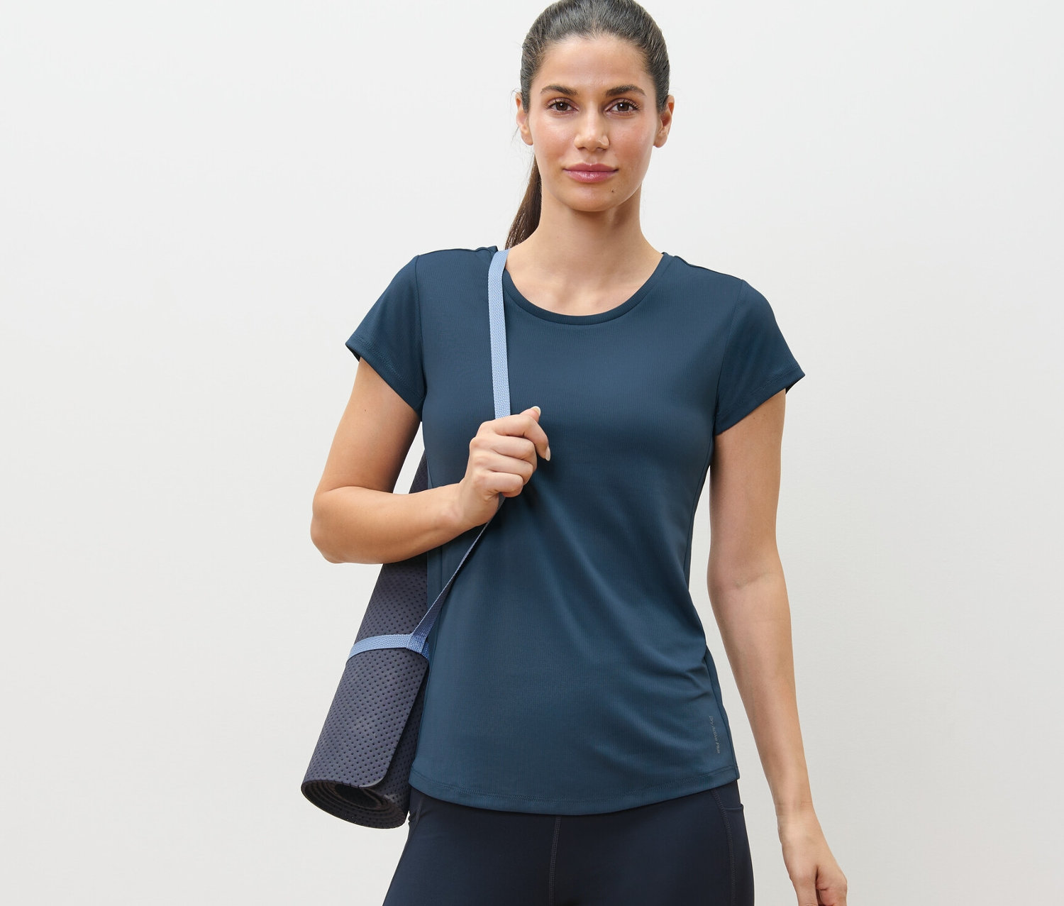 Kobieta ma na sobie niebieską koszulkę funkcyjną i niesie matę do ćwiczeń.