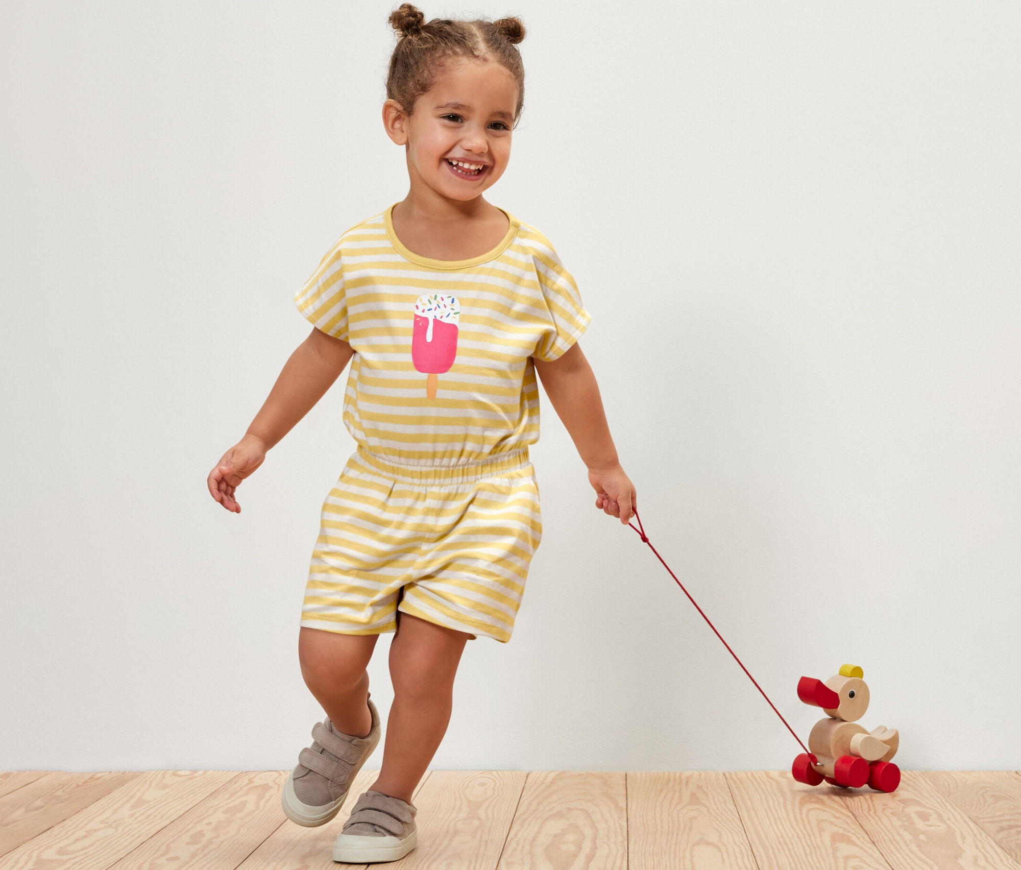 Uśmiechnięta dziewczynka w kombinezonie w paski ciągnie za sobą drewnianą zabawkę kaczkę.
