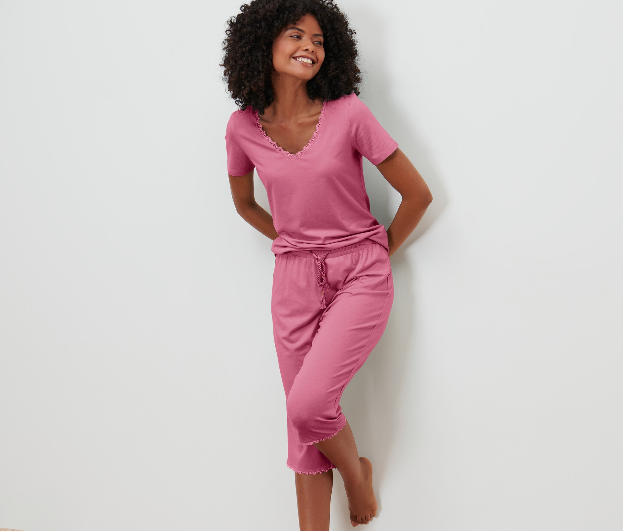 Uśmiechnięta kobieta opiera się o ścianę w różowej piżamie ze spodniami o długości 3/4.