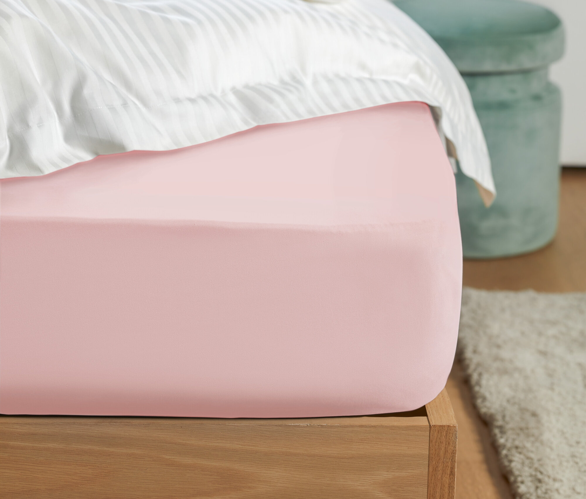 Zbliżenie na różowe prześcieradło z gumką na łóżku z białą pościelą w paski i zielonym stołkiem w tle.