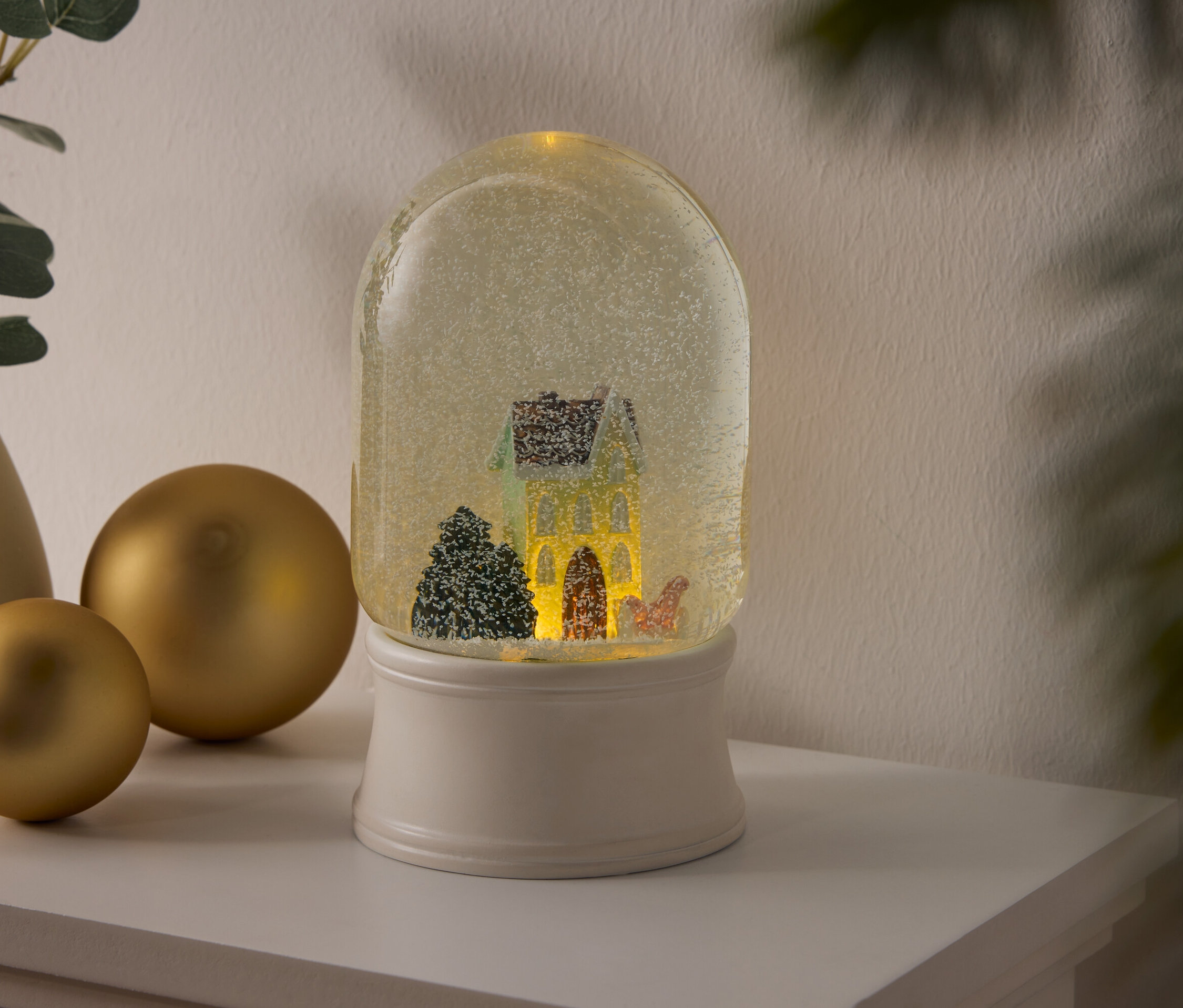 Kula śnieżna z domem i drzewami na białym cokole stoi na półce obok złotych kul i wazonu z roślinami.