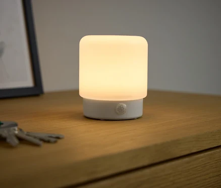 Na drewnianym stoliku nocnym świeci się lampka LED-Mini-Multifunktionslicht mit Bewegungsmelder.