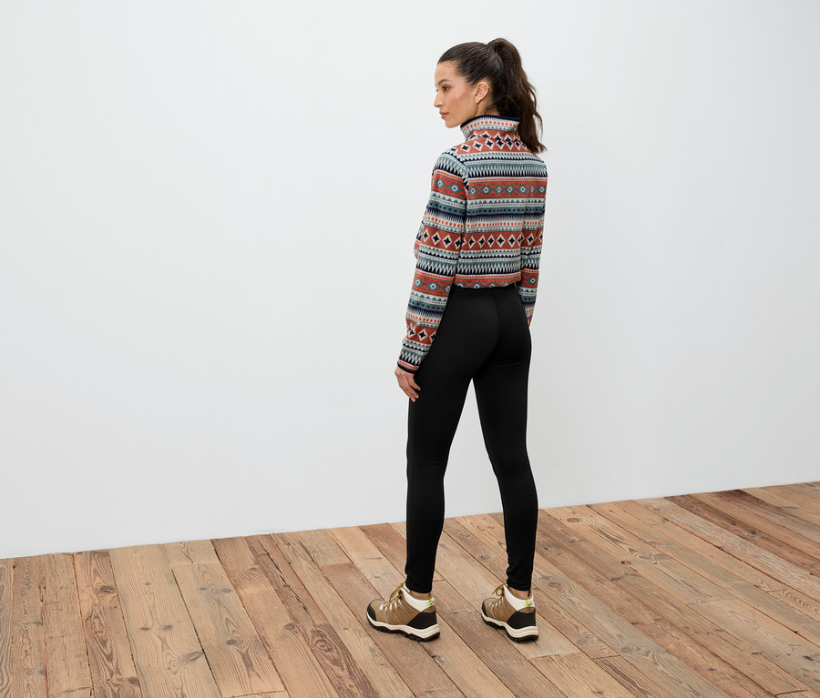 Kobieta z brązowymi włosami związanymi w kucyk stoi tyłem, ubrana w wzorzystą bluzkę z długim rękawem, czarne legginsy i brązowe buty.