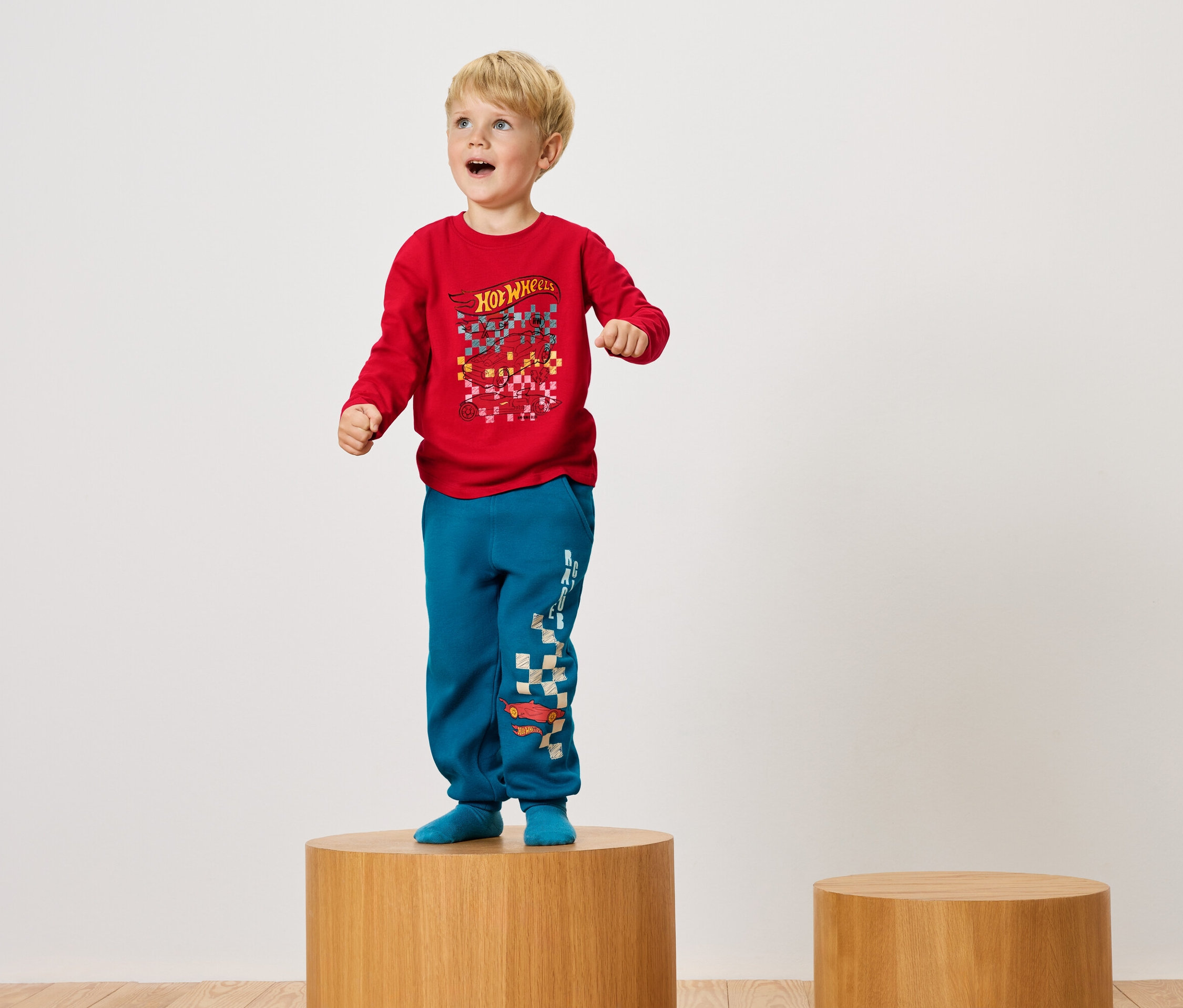 Chłopiec stoi na drewnianym postumencie w czerwonej koszulce i niebieskich spodniach.