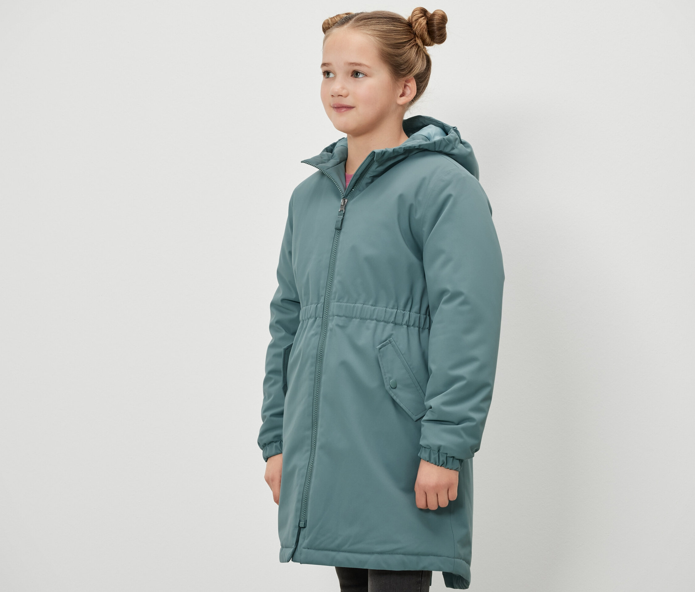 Dziewczyna stoi bokiem i ma na sobie zieloną parkę termiczną Kinder-Thermoparka.