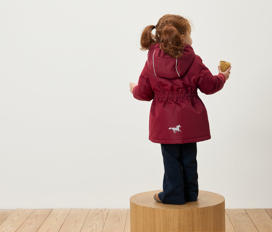 Ruda dziewczynka w kucykach stoi tyłem do aparatu na drewnianym postumencie, ma na sobie bordową kurtkę z kapturem i obrazkiem konia i trzyma w ręku wafelek.