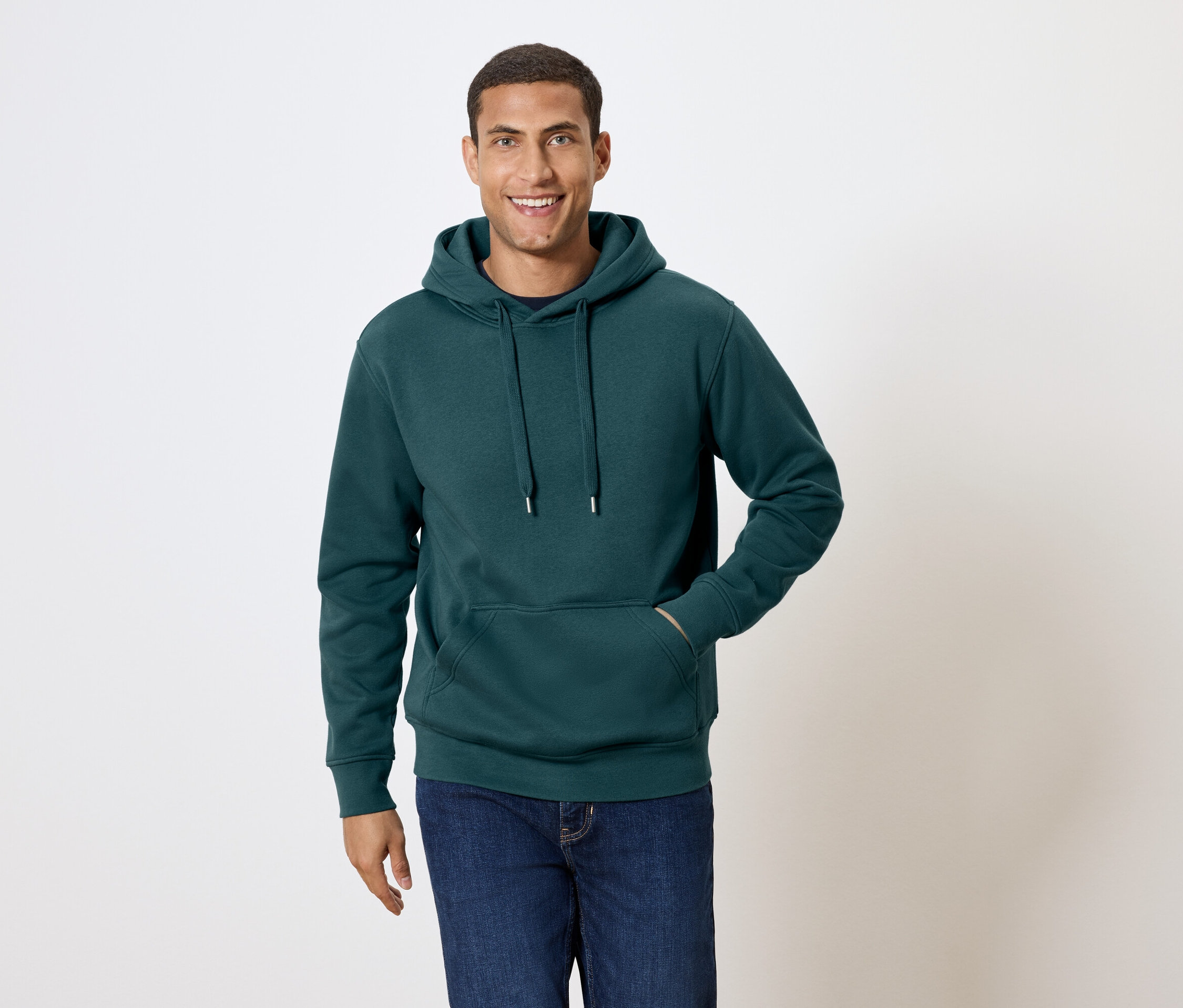 Mężczyzna z krótkimi włosami uśmiecha się i pozuje w ciemnozielonej bluzie z kapturem i niebieskich dżinsach.