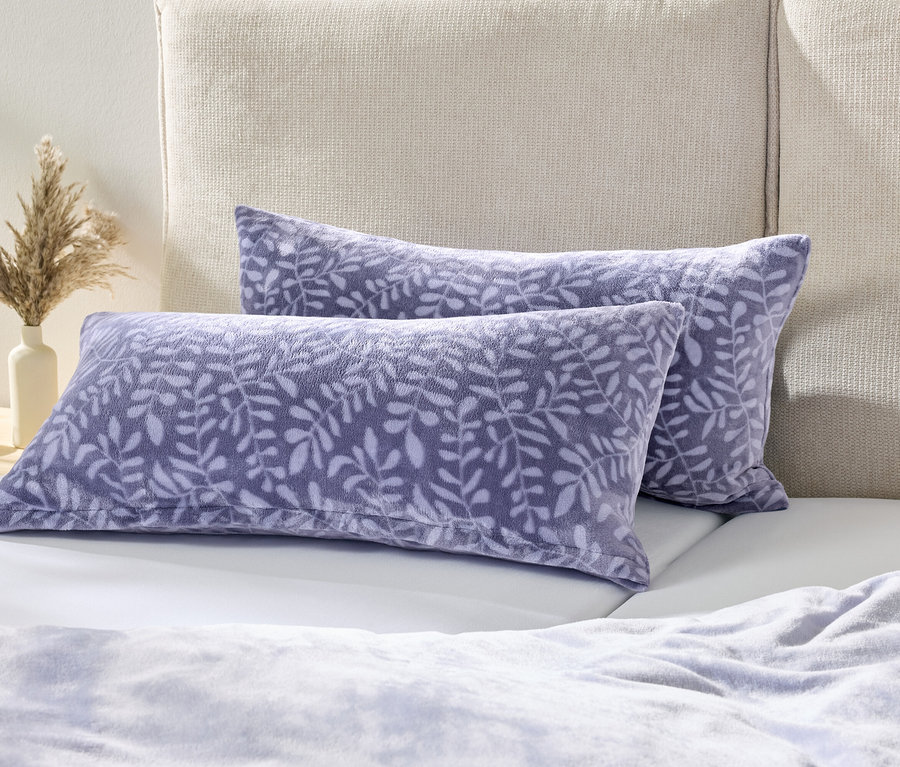 Dwie fioletowe poduszki z wzorem liści leżą na białym łóżku obok stolika nocnego z wazonem suszonych kwiatów.
