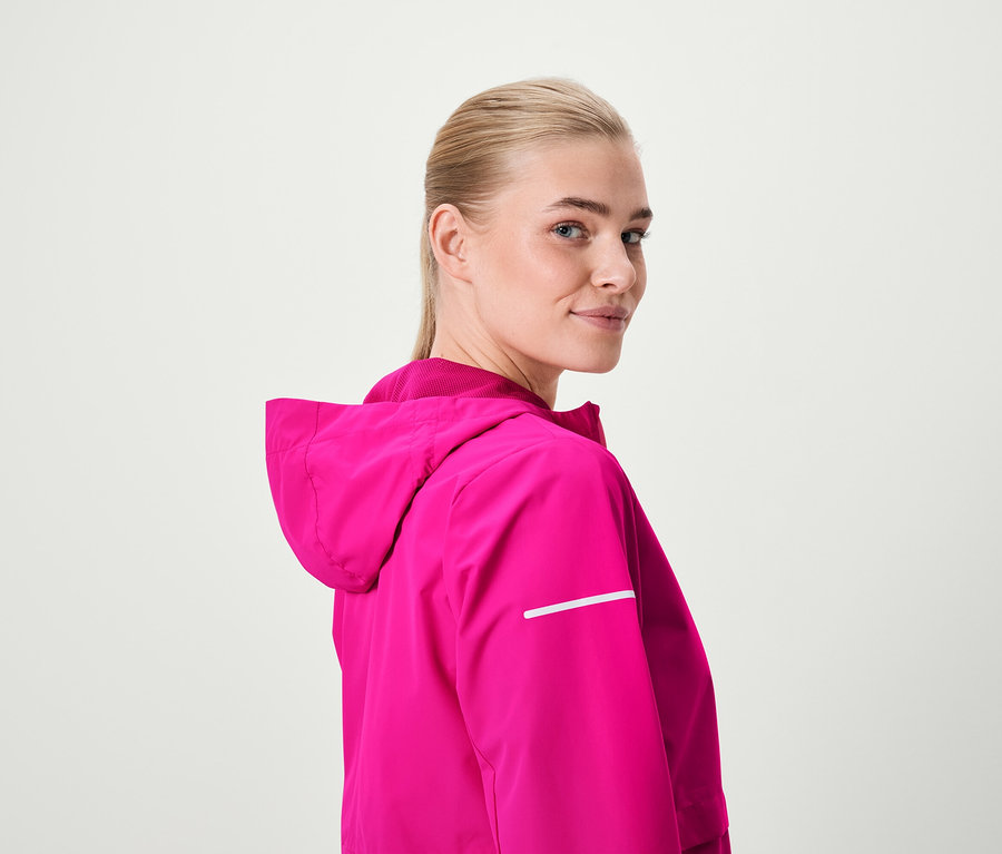 Blondynka spogląda przez ramię, ubrana w różową kurtkę z kapturem.