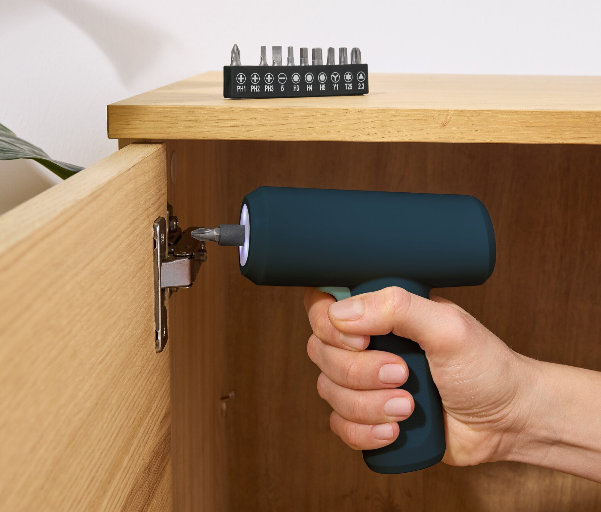Ręka trzyma niebieski mini wkrętak akumulatorowy 3,6 V i wkręca śrubę w drewnianą szafkę. Zestaw bitów leży na górze szafki.