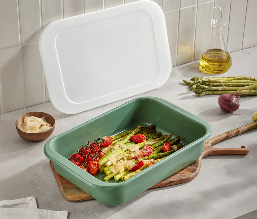 Zielona forma do pieczenia ze szparagami i pomidorami na drewnianym stole. Widoczna jest również biała pokrywka, oliwa z oliwek, parmezan i cebula.
