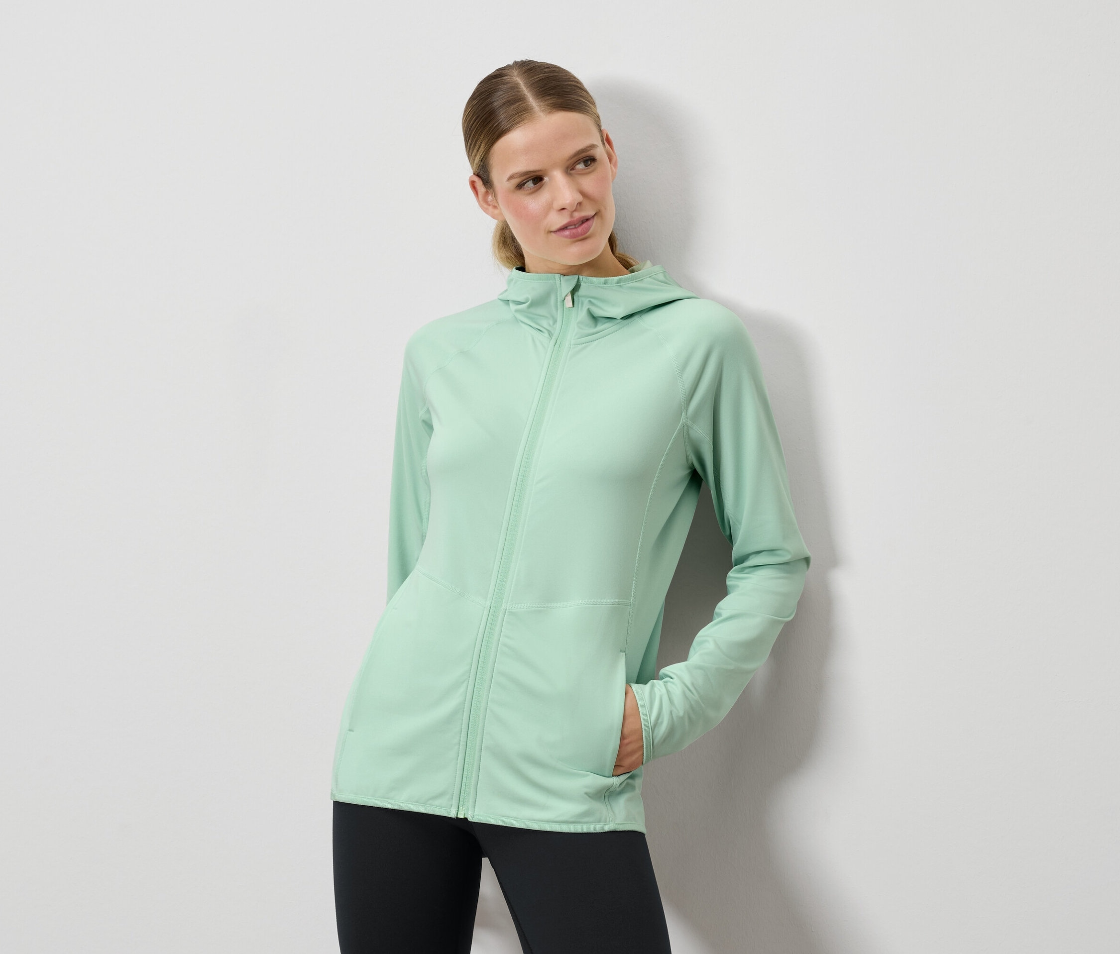 Kobieta pozuje w miętowo zielonej bluzie z kapturem i czarnych legginsach.