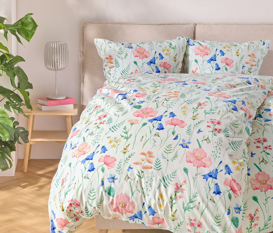 Łóżko z pościelą w kwiaty, obok stolik z lampą i książkami, po lewej roślina.