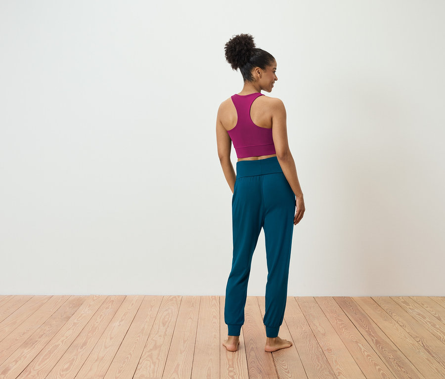 Kobieta stoi tyłem do kamery, ubrana w purpurowy biustonosz sportowy i niebiesko-zielone spodnie dresowe.