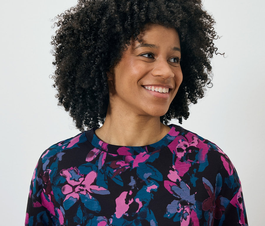 Portret uśmiechniętej kobiety z afro, ubranej w czarny top z kwiatowym wzorem w kolorze różowym i niebieskim.